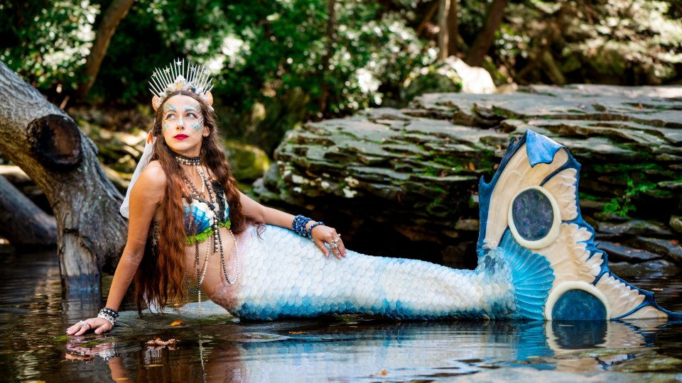 A mermaid in blue stares at the sky while sitting near the river's edge