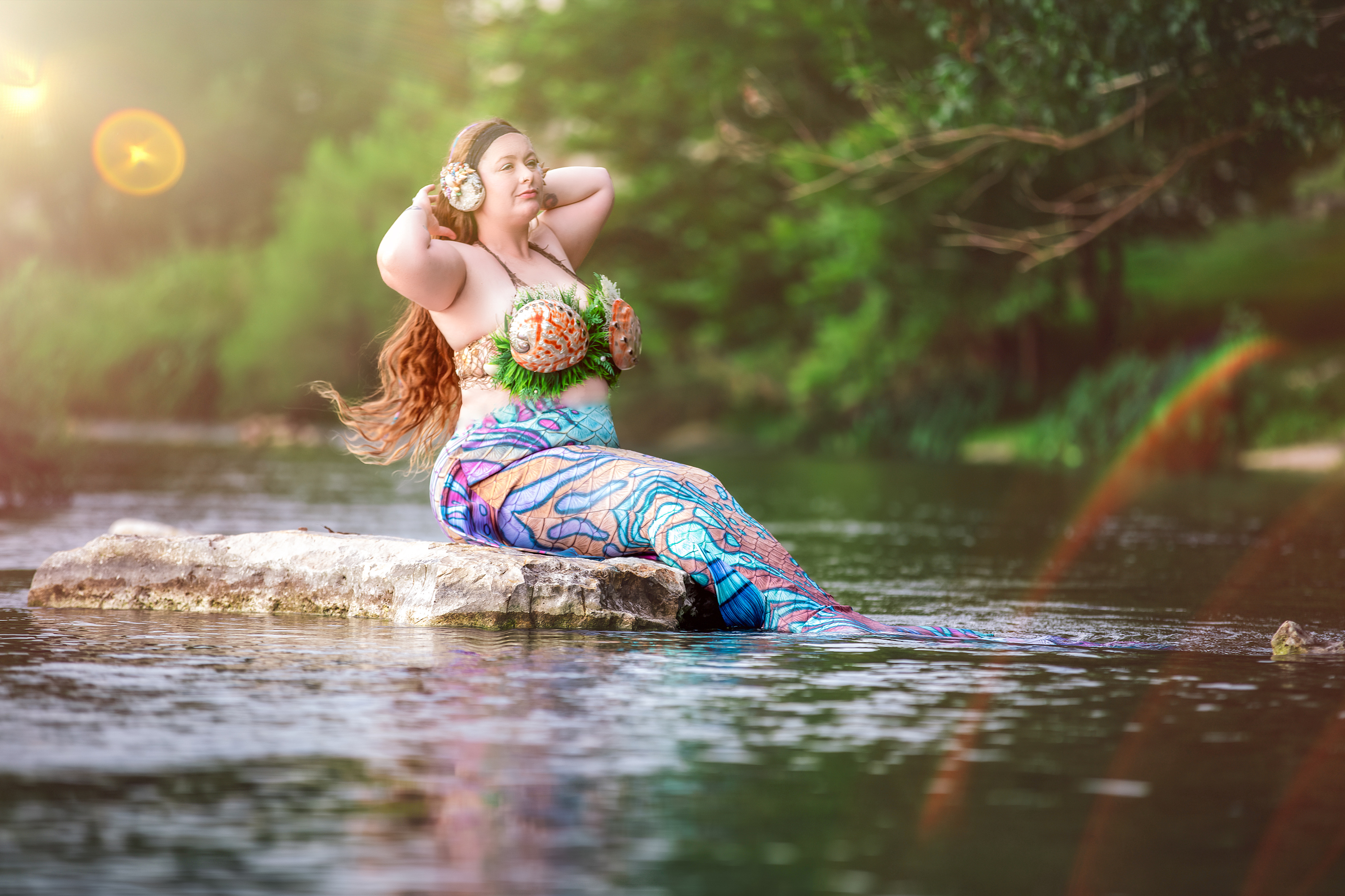 A colorful mermaid with long hair suns herself on a rock in a river